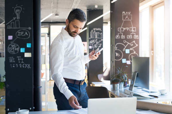 Handsome young businessman working in his office, standing at the desk, holding a smart phone, laptop in front of him.