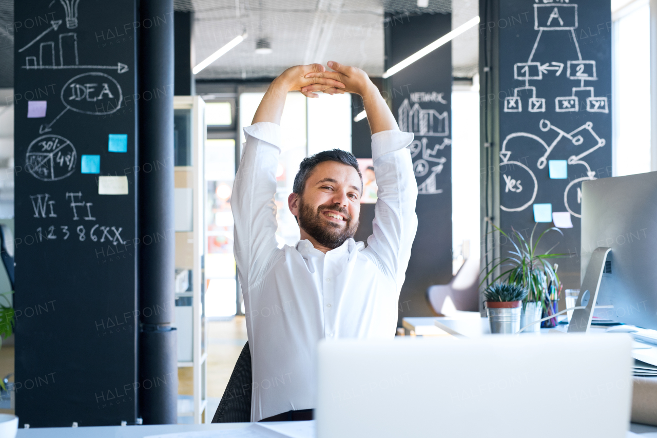 Handsome young businessman in his office, sitting at the desk, stretching his arms.