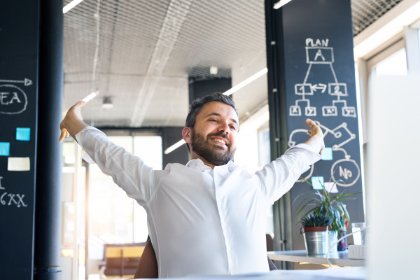 Handsome young businessman in his office, sitting at the desk, stretching his arms.