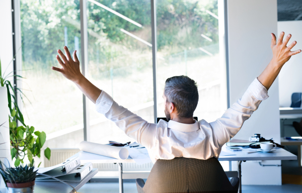 Handsome young businessman in his office, sitting at the desk, stretching arms. Rear view.