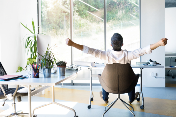 Handsome young businessman in his office, sitting at the desk, stretching his arms. Rear view.