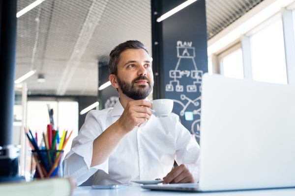 Handsome young businessman working in his office, sitting at the desk, laptop in front of him, drinking coffee.