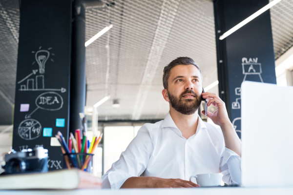 Young hipster businessman working in his office, sitting at the desk, holding coffee and smart phone, making phone call.