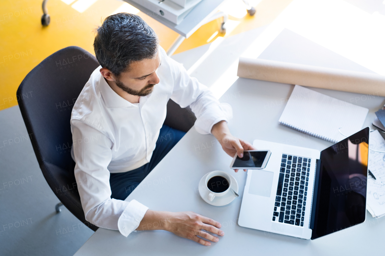 Handsome young businessman working in his office, sitting at the desk, holding a smart phone, laptop and coffee in front of him.