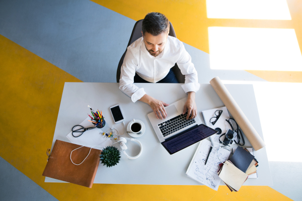 Young hipster businessman in his office, sitting at the desk, working, laptop in front of him. High angle view.