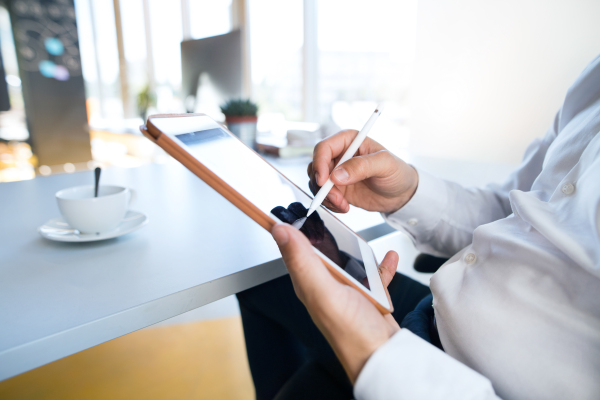 Unrecognizable businessman in his office, sitting at the desk, working on tablet, coffee cup in front of him.