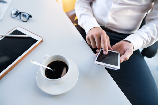 Unrecognizable young businessman working in his office, sitting at the desk, holding a smart phone, tablet and coffee in front of him.