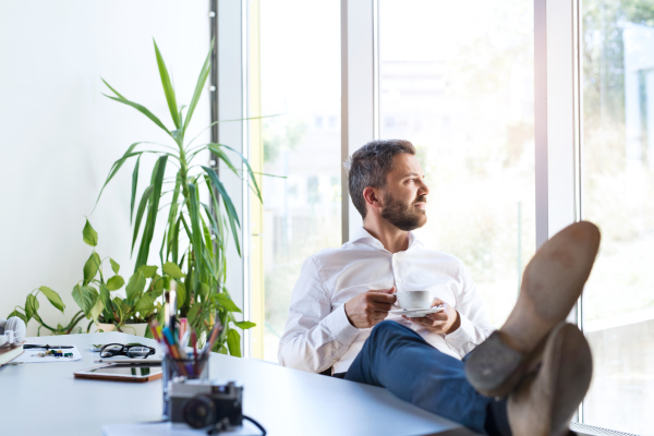 Handsome young businessman working in his office, sitting at the desk, laptop in front of him, drinking coffee.