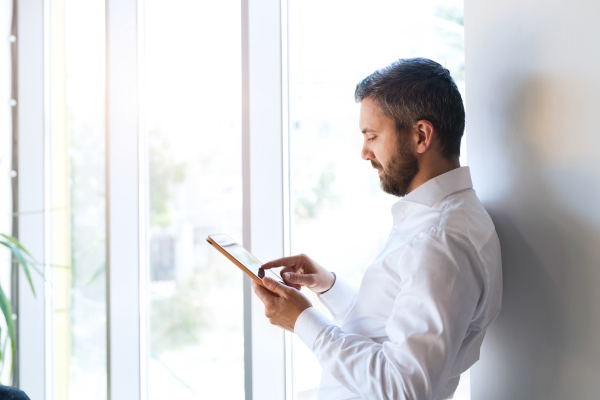 Hipster businessman in his office, standing at the window, working on tablet.