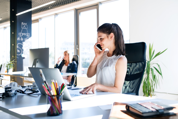 Two attractive young business women in the office working, one of them holding a smart phone, making a phone call.