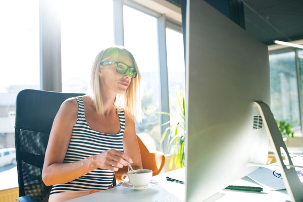 Beautiful young businesswoman with computer in her office, sitting at the desk, working and drinking coffee..