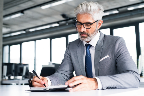 Mature businessman in gray suit sitting at desk in the office, writing something into his personal organizer.