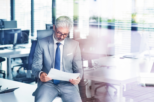 Mature businessman in gray suit sitting at desk in the office, reading documents.