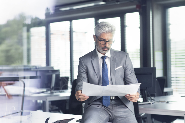 Mature businessman in gray suit sitting at desk in the office, reading documents.