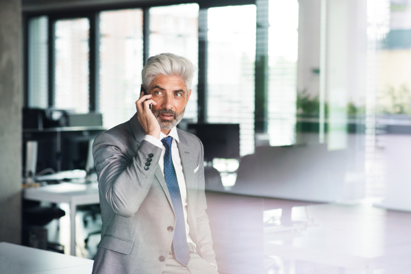 Mature businessman in gray suit in the office standing at the window holding smartphone making phone call. Shot through glass.