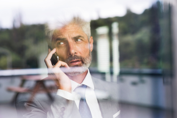 Mature businessman in gray suit in the office standing at the window holding smartphone making phone call. Shot through glass. Close up.