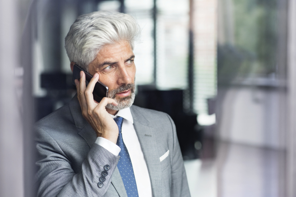 Mature businessman in gray suit in the office standing at the window holding smartphone making phone call.