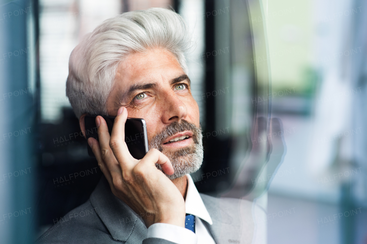 Mature businessman in gray suit in the office standing at the window, holding a smartphone, making a phone call.