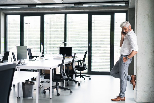 Mature businessman in white shirt in the office standing, holding smartphone and making phone call.