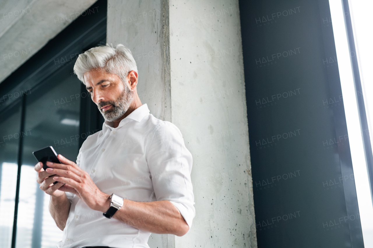 Mature businessman in white shirt in the office standing at the concrete wall, holding a smartphone, texting.