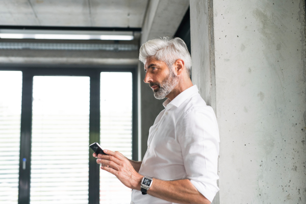 Mature businessman in white shirt in the office standing at the concrete wall, holding a smartphone, texting.