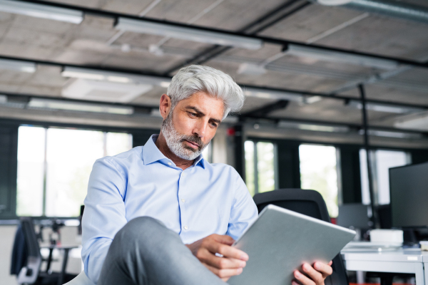 Mature businessman with tablet at the desk in the office, reading documents.