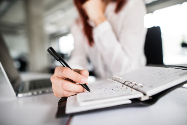 Unrecognizable businesswoman in the office, sitting at the desk, writing something into her personal organizer. Close up.