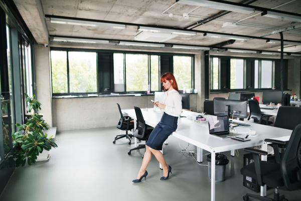 Beautiful young businesswoman with smartphone in her office sitting on desk, having a break.