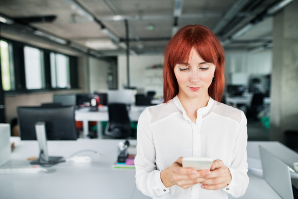 Beautiful young businesswoman with smart phone in her office, texting.