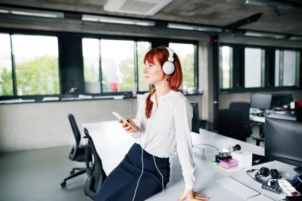 Beautiful young businesswoman in her office wearing big white headphones, holding a smart phone, listening music.