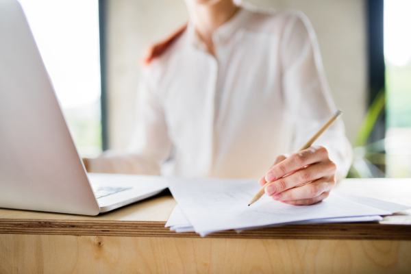 Unrecognizable young businesswoman in her office, sitting at the desk, laptop in front of her, writing something on paper.