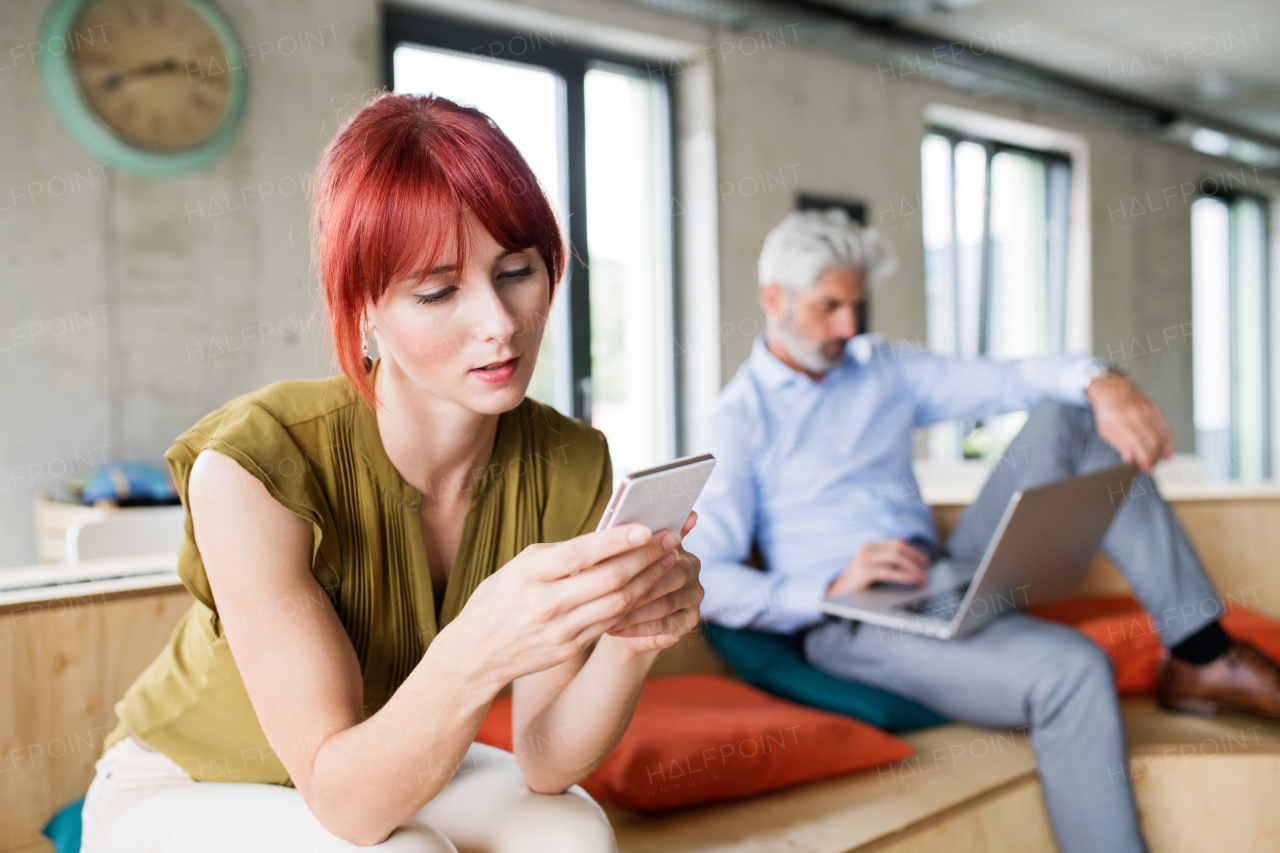 Two business people in creative office working. Woman holding a smart phone, texting. Man working on laptop.