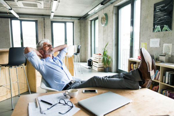 Thoughtful mature businessman with gray hair in the office with feet up on desk. Man with smartphone and laptop on the desk.