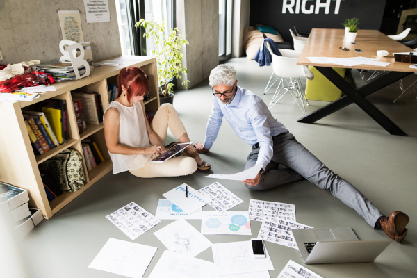 Two business people with tablet in the workplace. Woman and man sitting on the floor in the office consulting a project together.