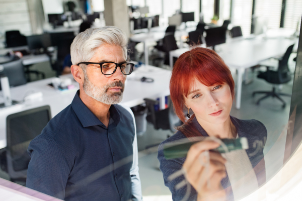 Handsome mature businessman and young businesswoman in the office writing notes, setting goals. Shot through glass.