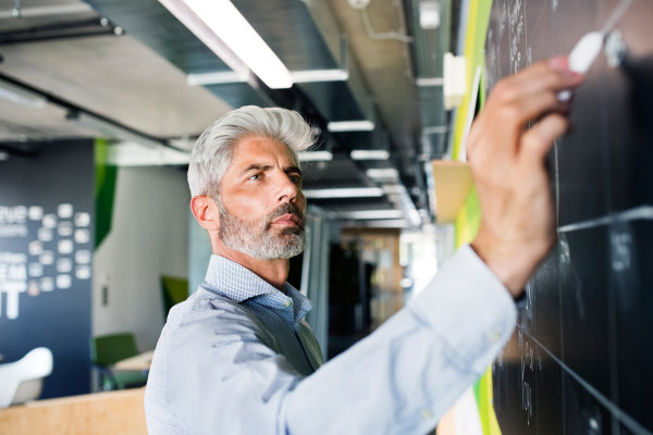 Handsome mature businessman with gray hair in the office writing notes on blackboard, setting goals.