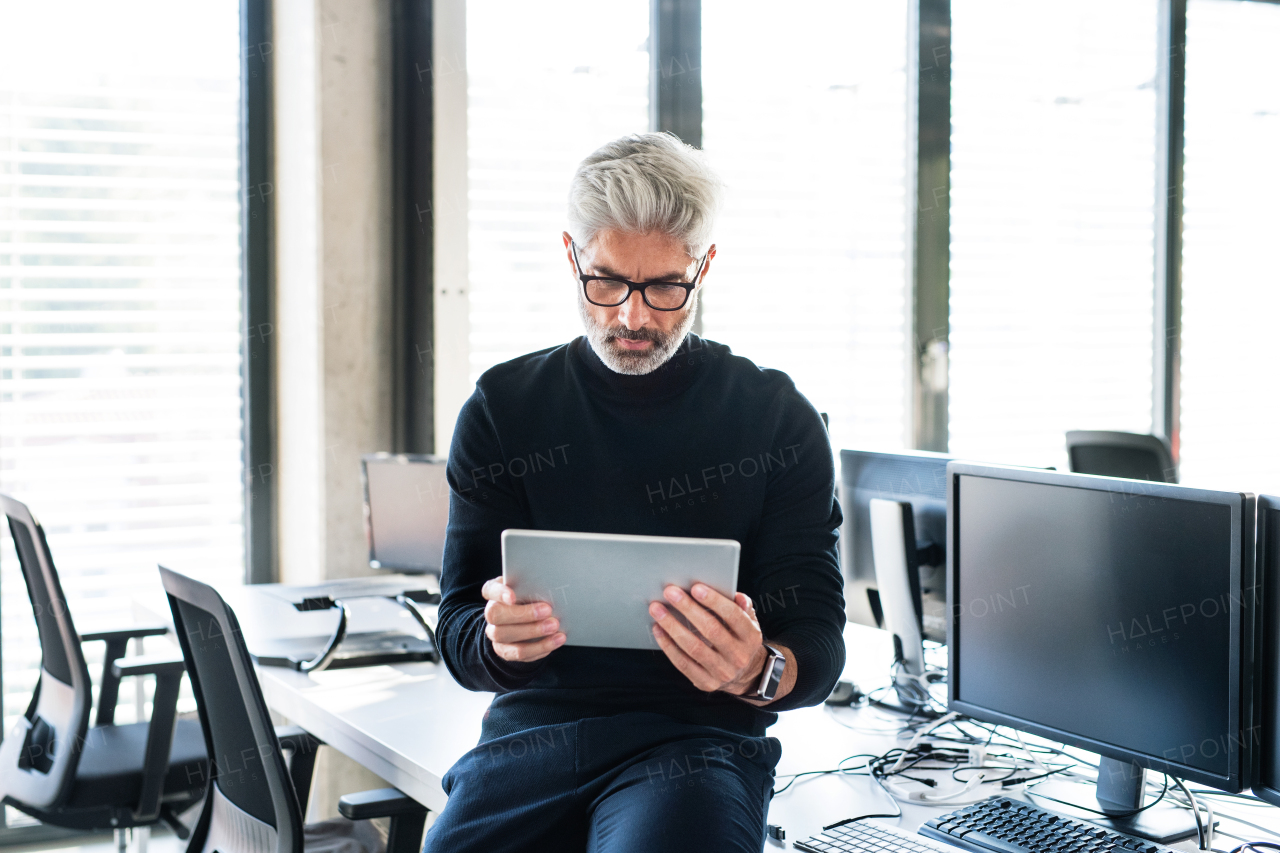 Handsome mature businessman with tablet in the office working, reading or searching something. Man sitting on the desk.