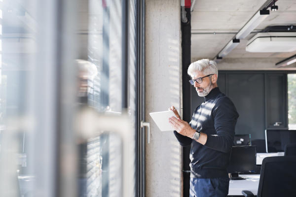 Mature businessman with tablet at the window in the office working, reading documents.