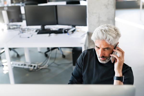 Mature businessman with smart phone sitting at the desk in the office, making a phone call, computer in front of him.