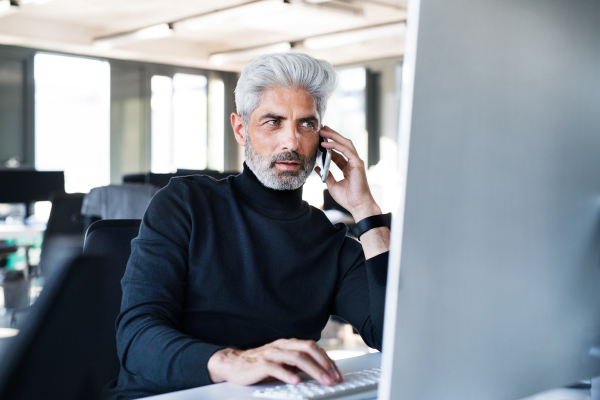 Mature businessman with laptop and smart phone sitting at the desk in the office, making a phone call.