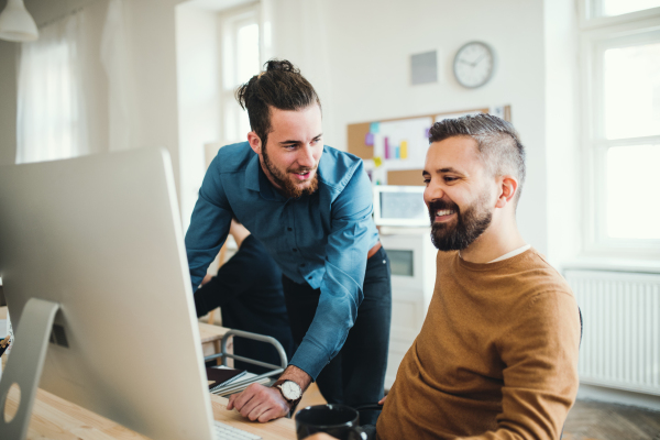 Two young hipster male businesspeople with computer talking in a modern office.