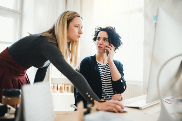 Two female business people with smartphone and computer in a modern office, talking.