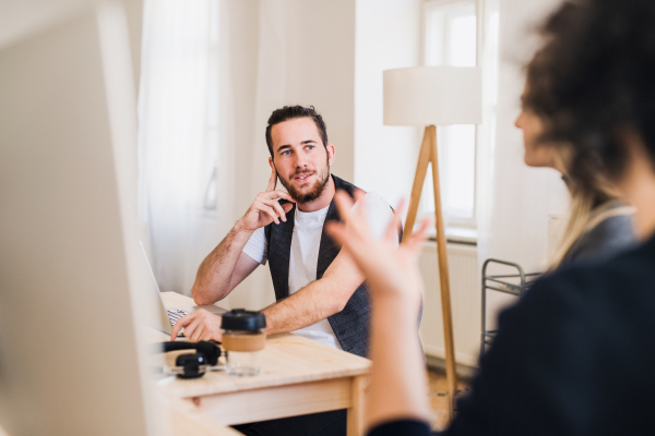 A portrait of young hipster businessman in a modern office, talking to unrecognizable colleagues.