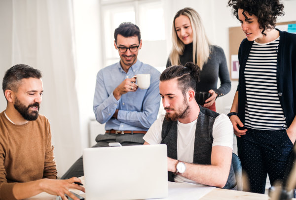 Group of young, cheerful businesspeople with laptop sitting and standing around table in a modern office, having meeting.