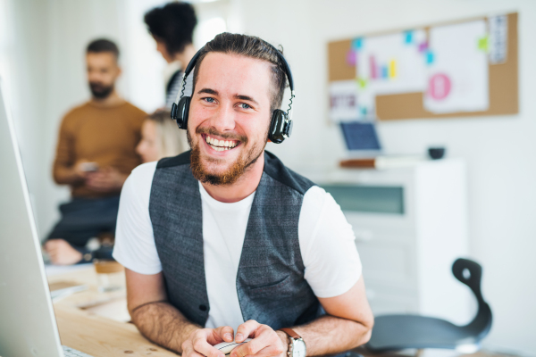 A portrait of young hipster businessman with headphones and colleagues in a modern office, laughing.