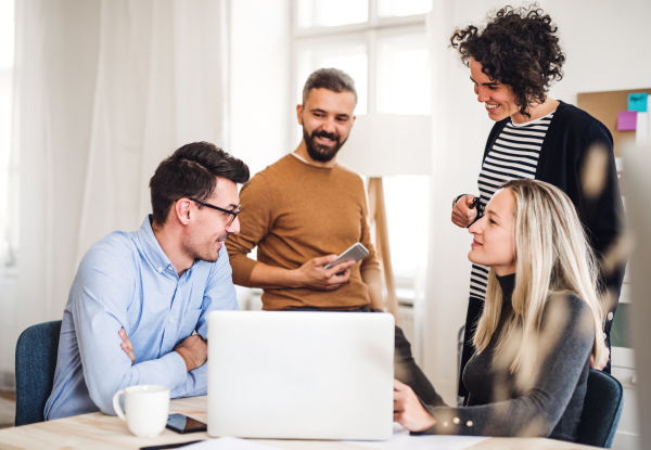 Group of young, cheerful businesspeople with laptop sitting and standing around table in a modern office, having meeting.