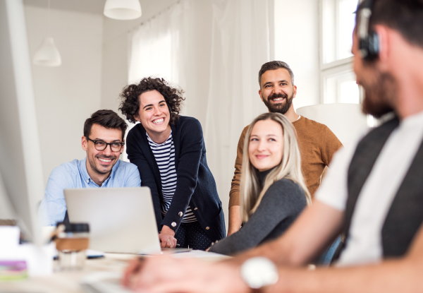 Group of young, cheerful businesspeople with laptop sitting and standing around table in a modern office, having meeting.