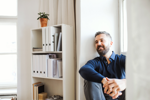 Mature hipster businessman sitting on a window sill in a modern office, resting. Copy space.