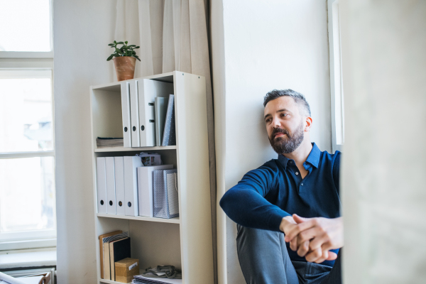 Serious mature hipster businessman sitting on a window sill in a modern office, resting. Copy space.