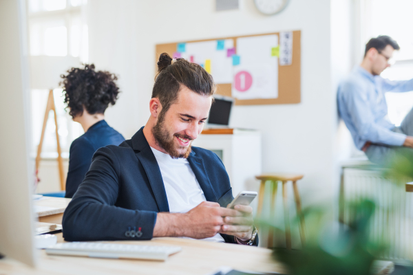 A portrait of young hipster businessman with smartphone and colleagues in the background in a modern office.